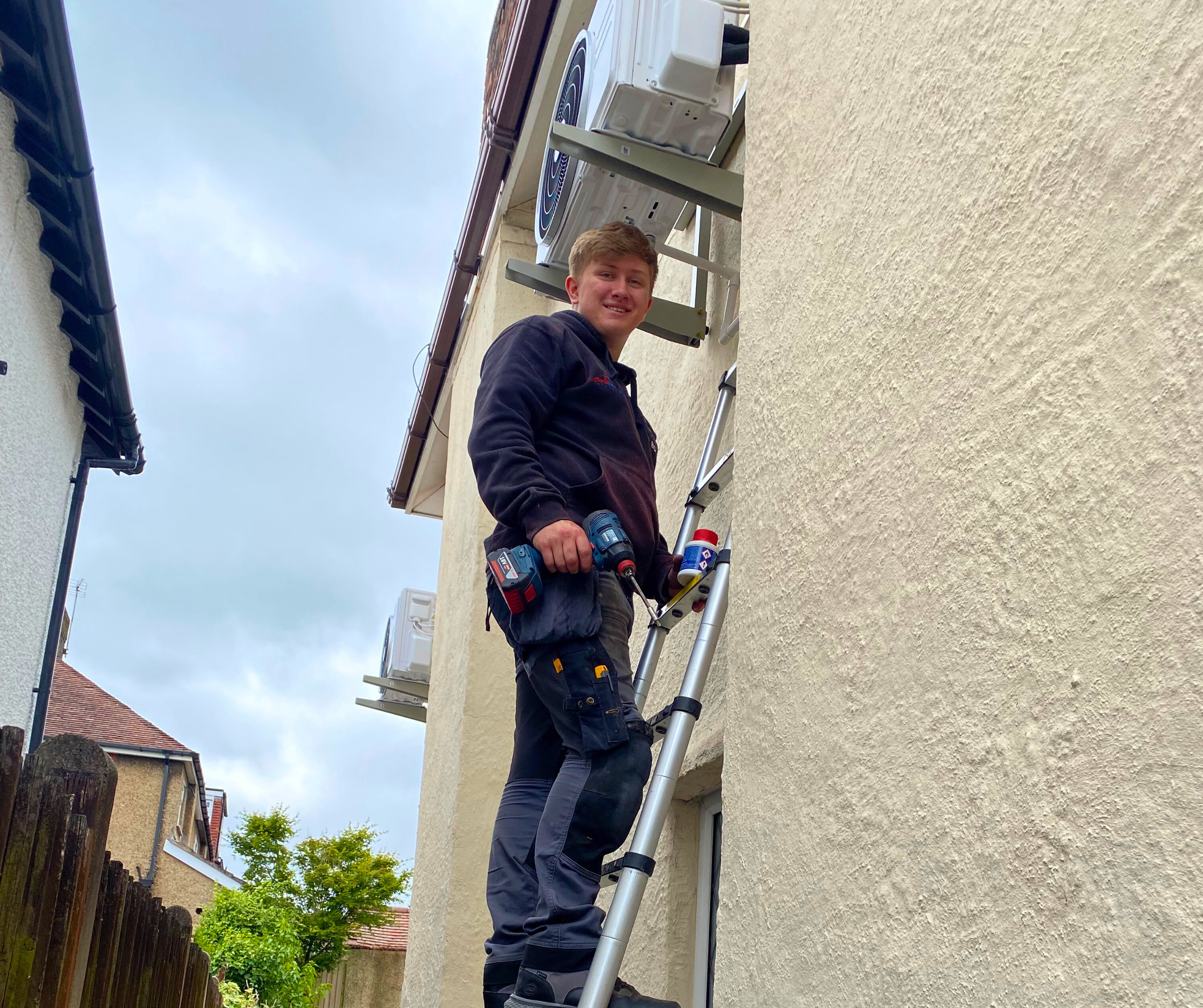 A young gas engineer from Home Heating Systems installing an outdoor air conditioning unit on the Wirral, working up a ladder with tools in hand.